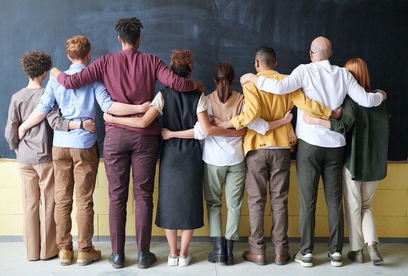 Eight people standing next to each other, backs to the camera, with their arms around each others' shoulders.