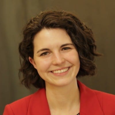 Headshot of Bridget Thoreson, a white person with wavy brown chin-length hair, wearing a red suit jacket and smiling at the camera