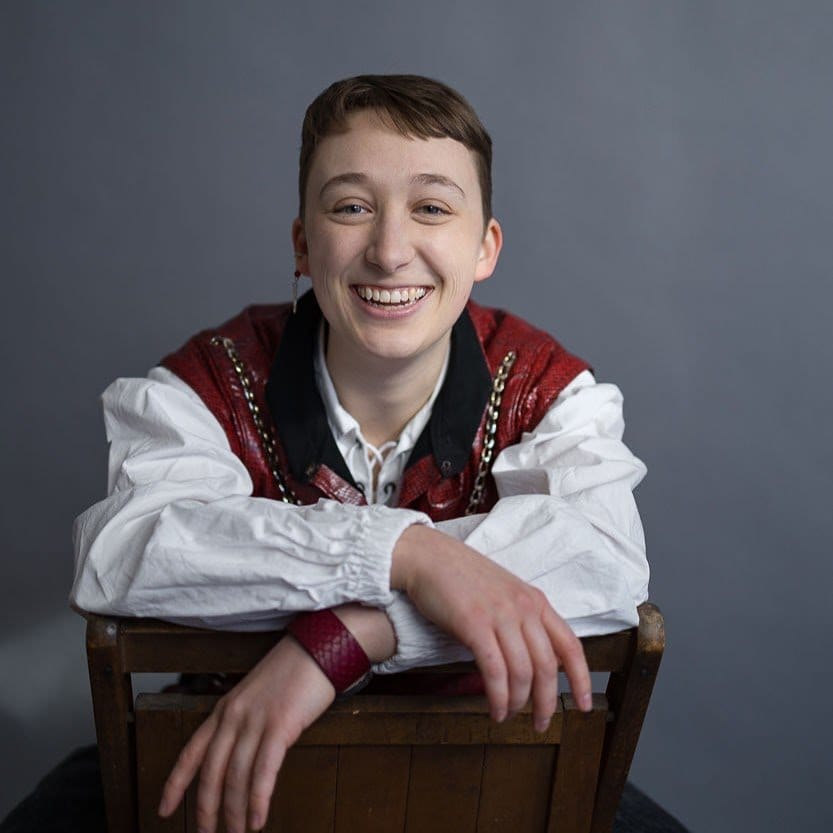 A photo of Beks Roen, a light-skinned person with short hair smiling at the camera, wearing a historical-looking white shirt and embroidered vest