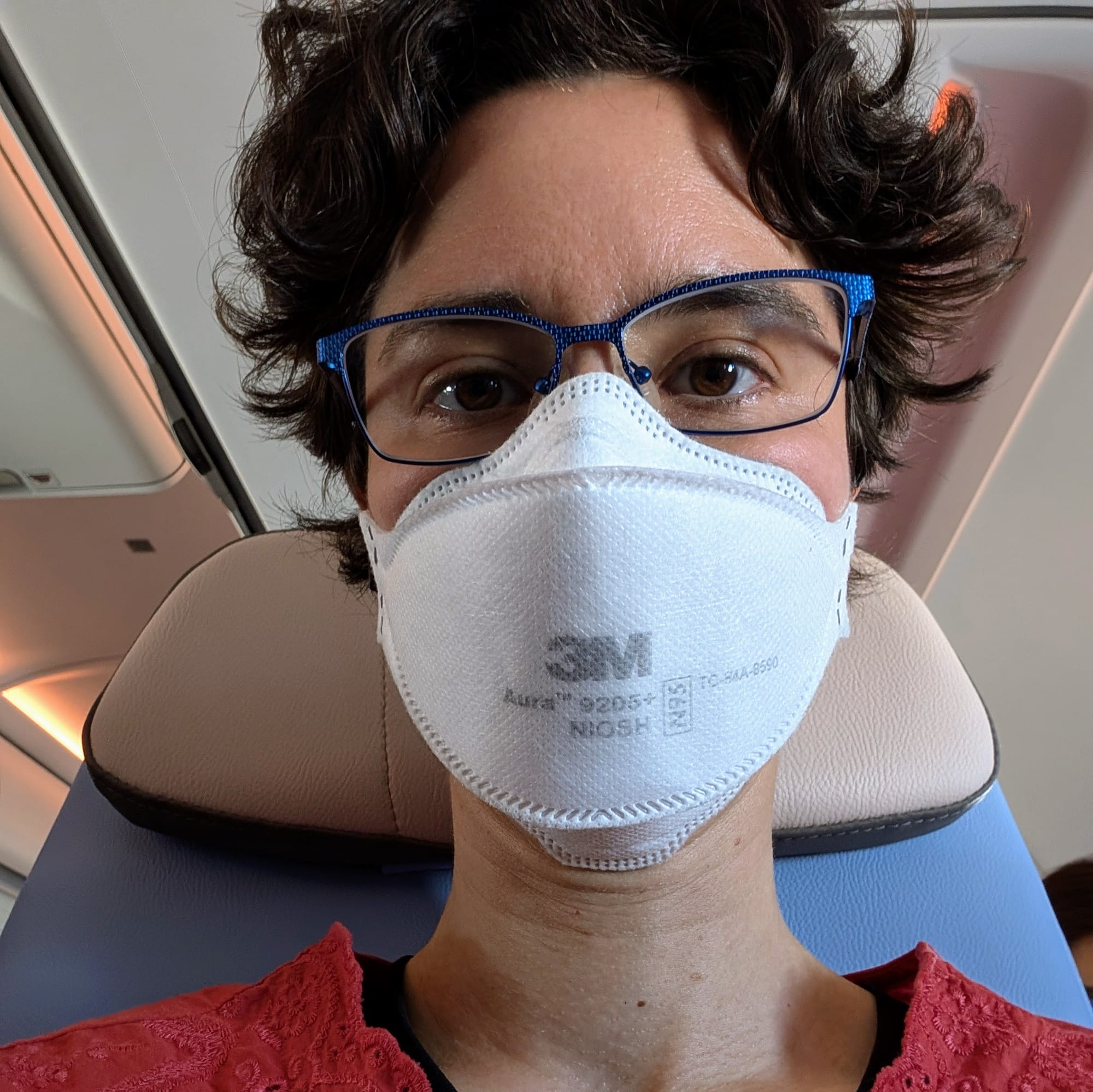Rey, a white non-binary person with short, curly brown hair, wearing glasses and an N95 mask, sitting in an airplane seat