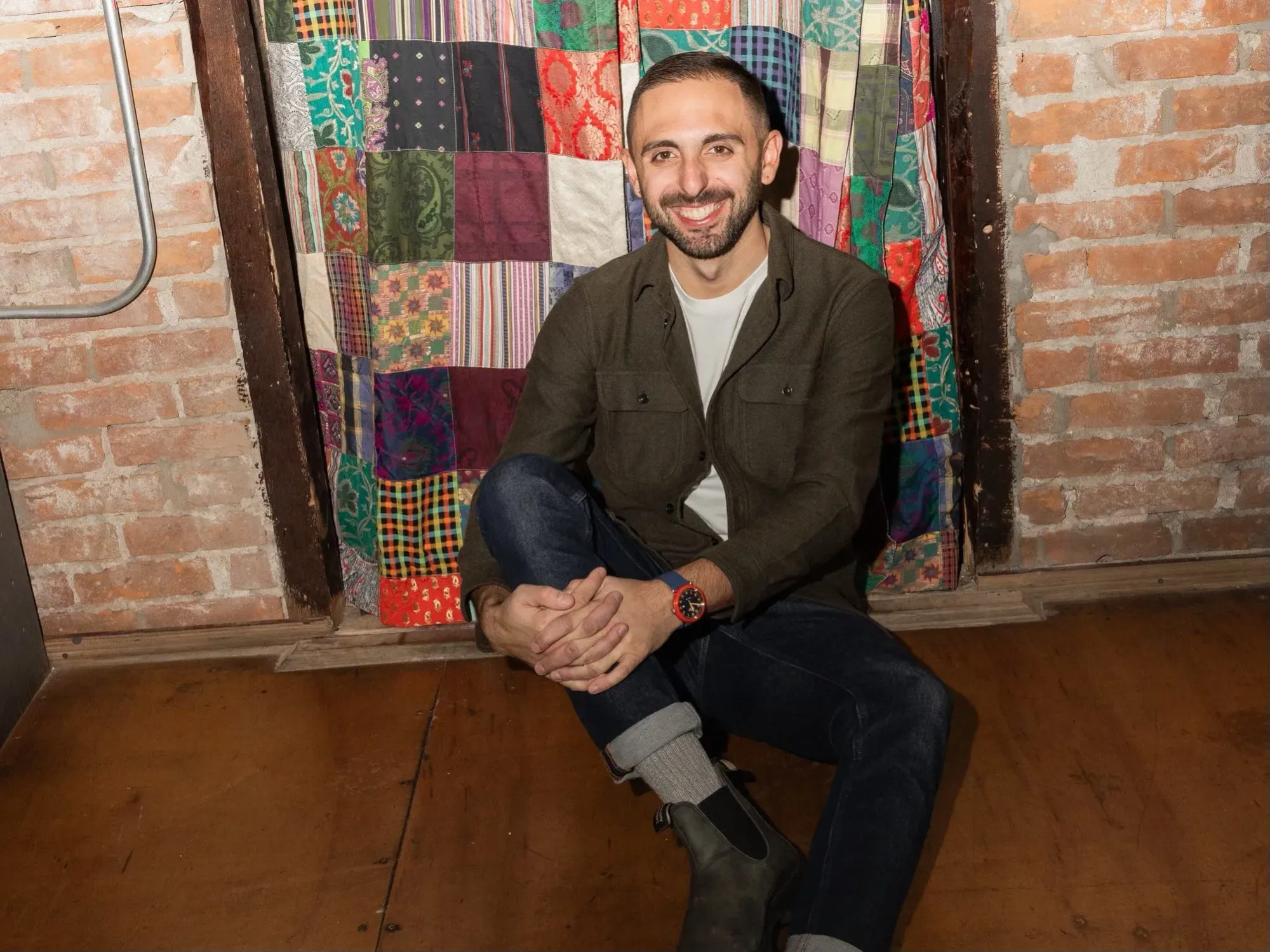 Photo of Mike sitting on the floor in front of a quilt, smiling, with short brown hair and a beard