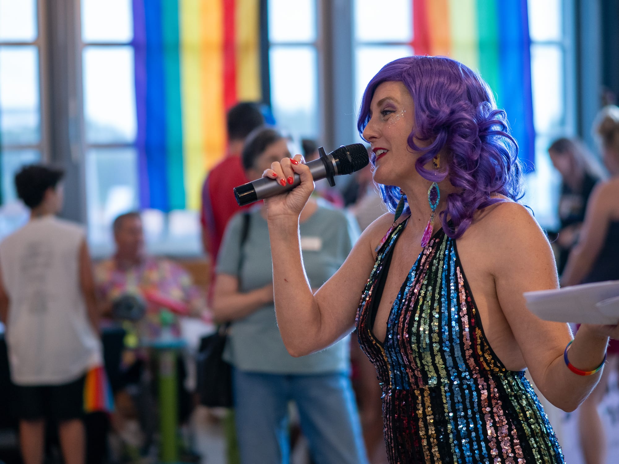 A white person wearing a purple wig and sequin rainbow dress speaks into a mic at an event, with people standing informally behind and rainbow flags hanging near the windows