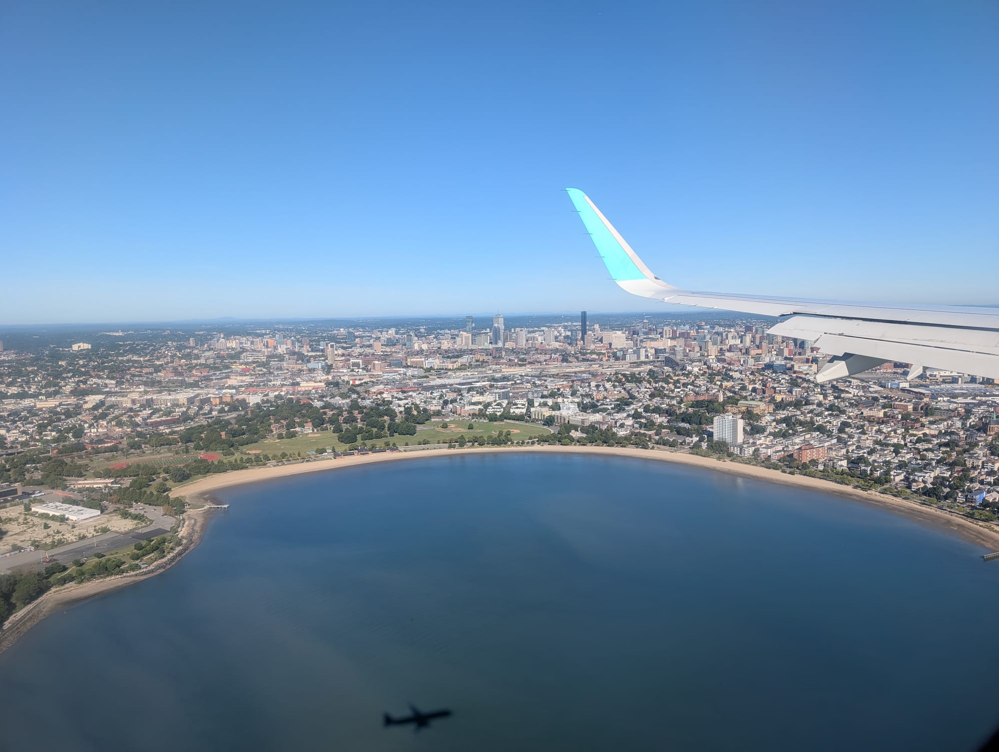 A photo taken out the window of a plane, of a circular body of water with the city in the background, and the shadow of the plane on the water